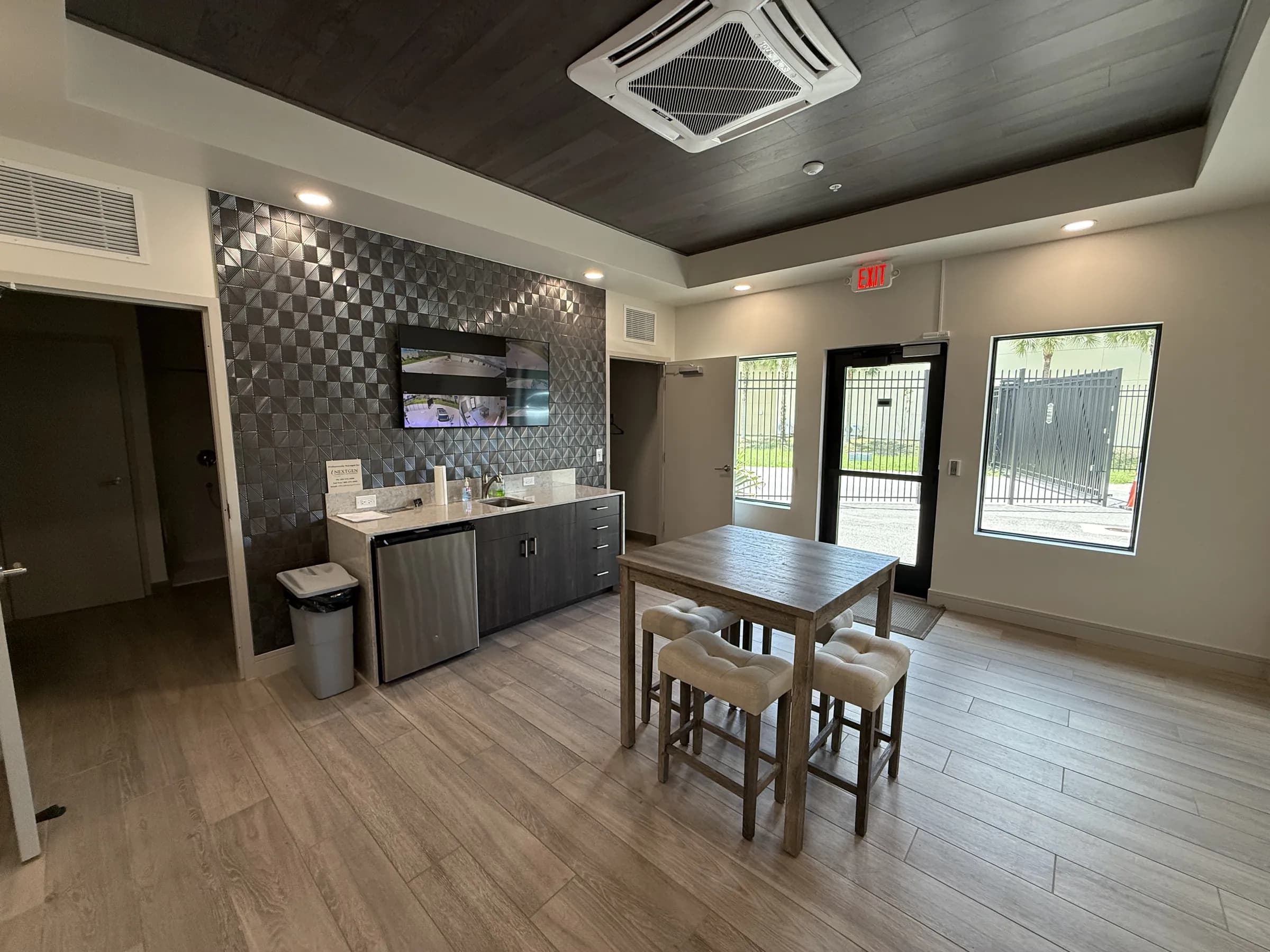 Wide view of the common room — high-top dining table and stools, kitchenette, wood-plank tray ceiling and glass entry doors.