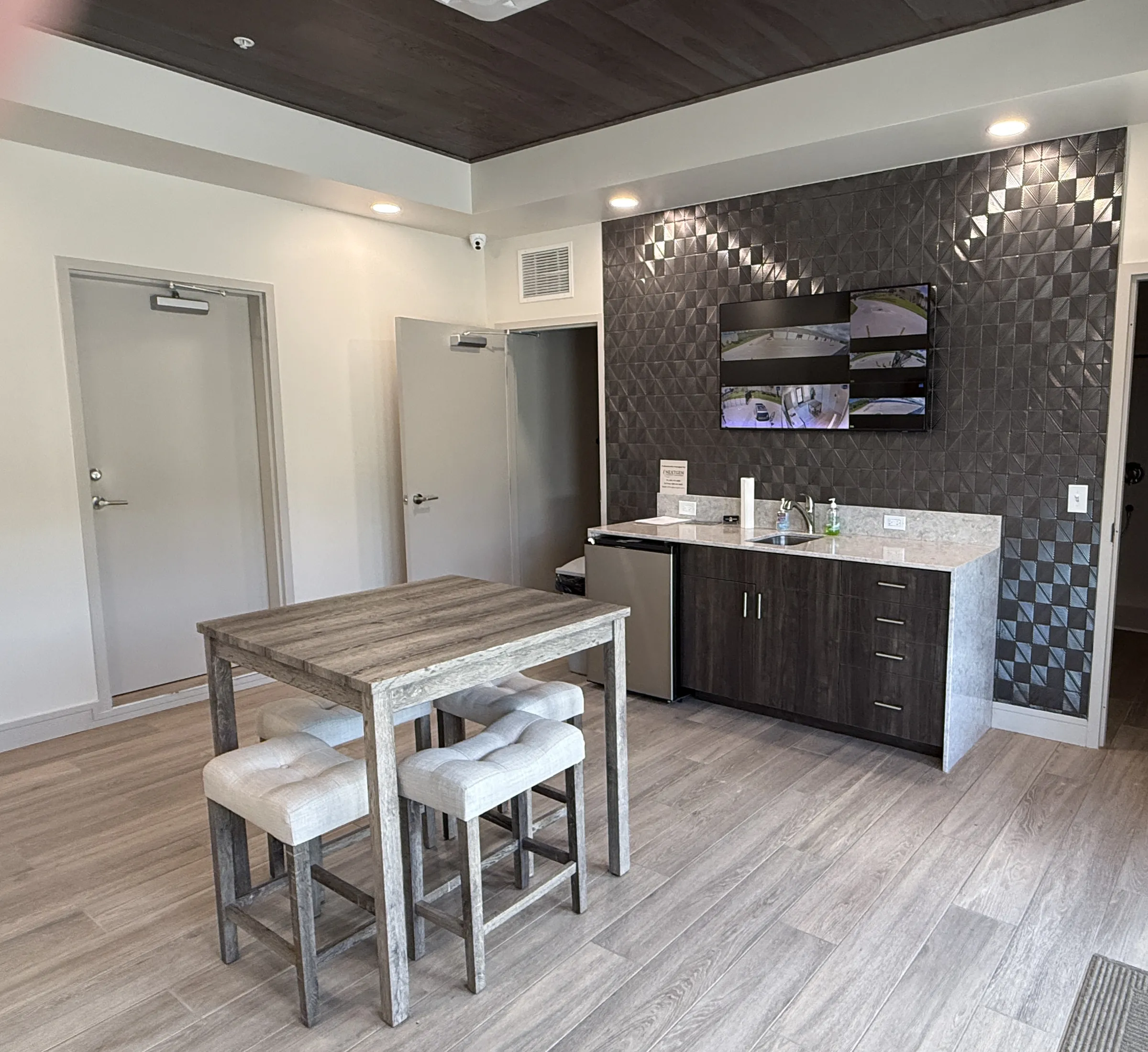 Common room kitchenette with quartz counter, dark cabinets, stainless mini fridge and a geometric tile accent wall below a mounted display.