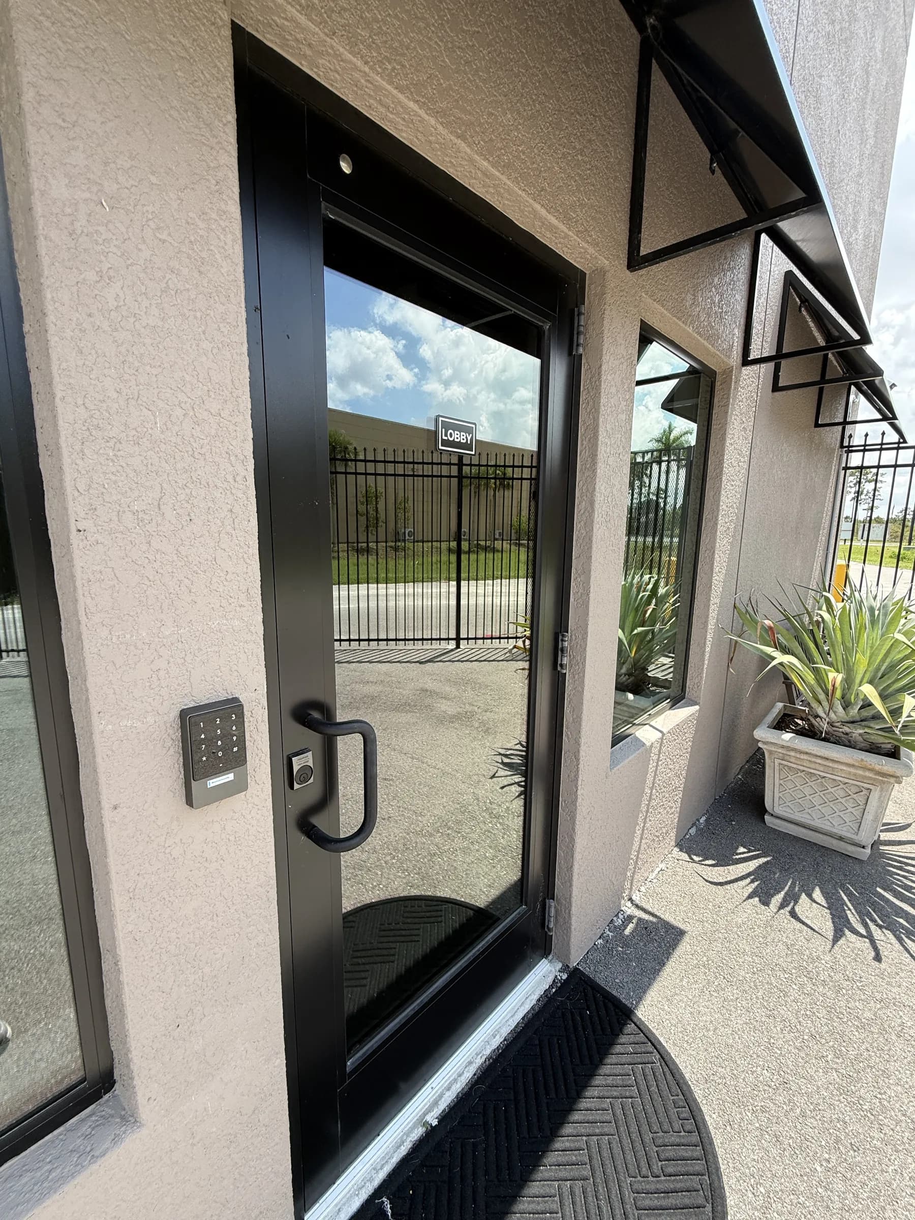 The glass lobby door with its black frame, wall-mounted keypad and fob reader, awnings and planter beyond.