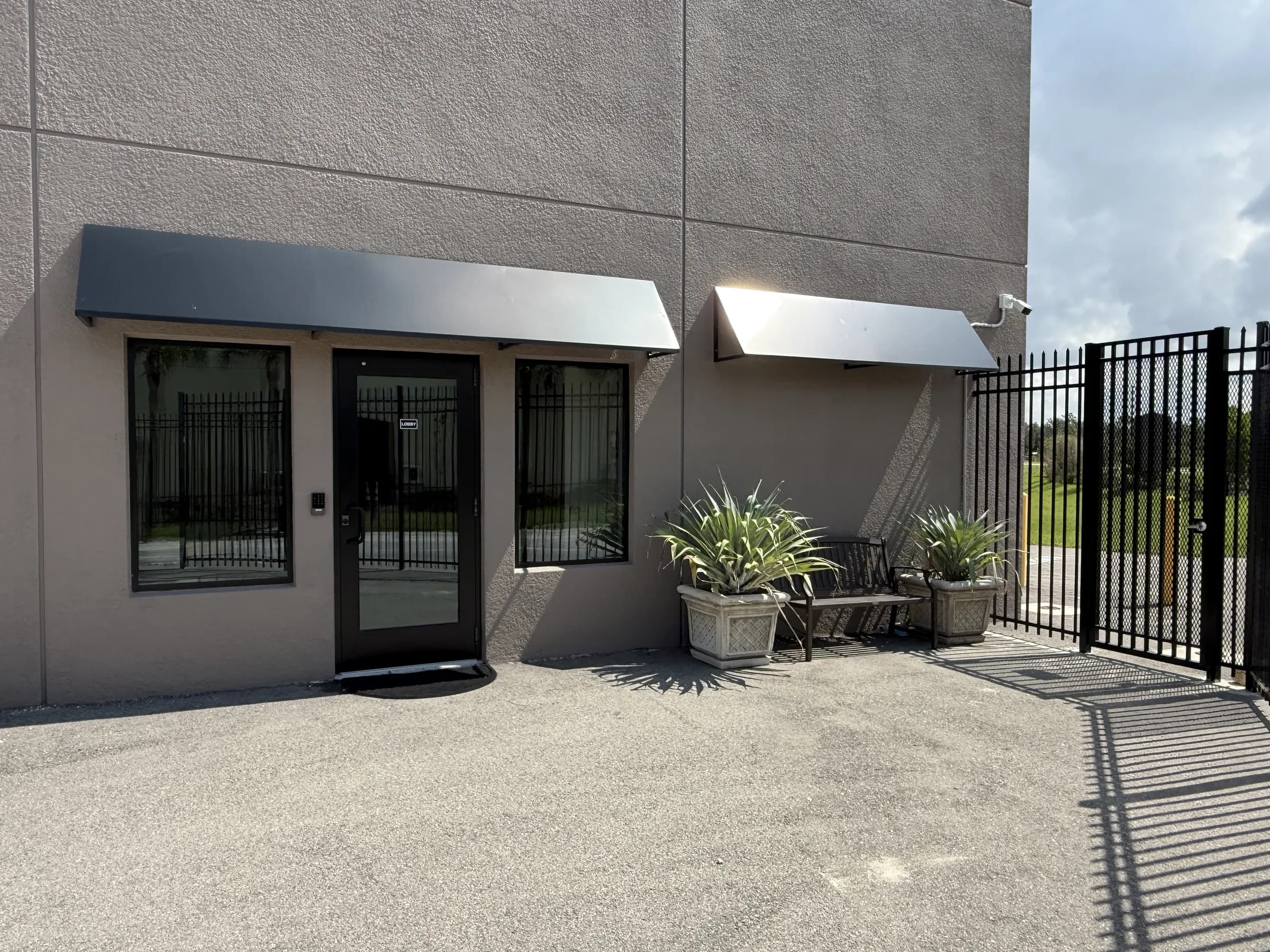 Exterior of the common room lobby entry — metal awnings, glass door, potted agaves and a bench in midday sun.