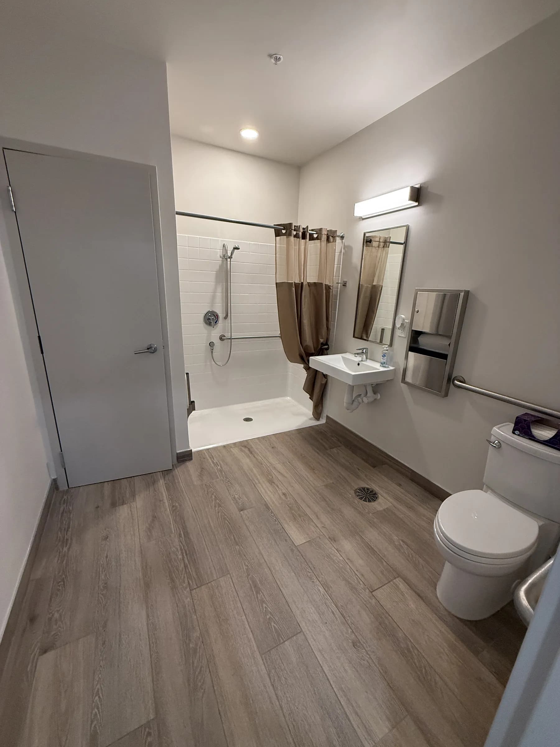 Common restroom with a curbless tiled shower, wall-hung sink, mirror and grab bars over wood-look plank flooring.
