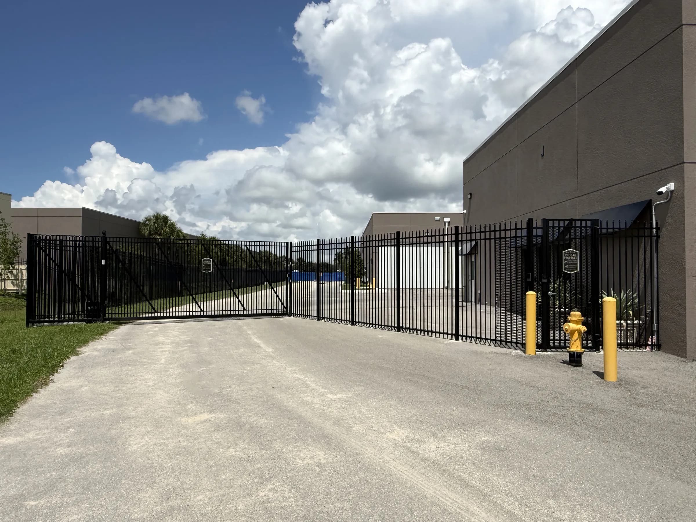 The black cantilever slide gate closed across the Vehicle Fortress entry lane, building wall and yellow bollards to the right under towering clouds.
