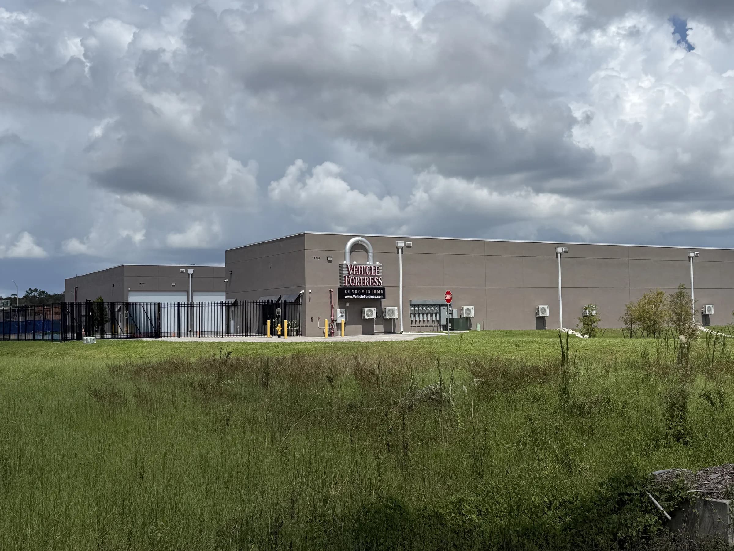 Distant view across an open field toward the Vehicle Fortress frontage, entry gate and sign under heavy gray storm clouds.