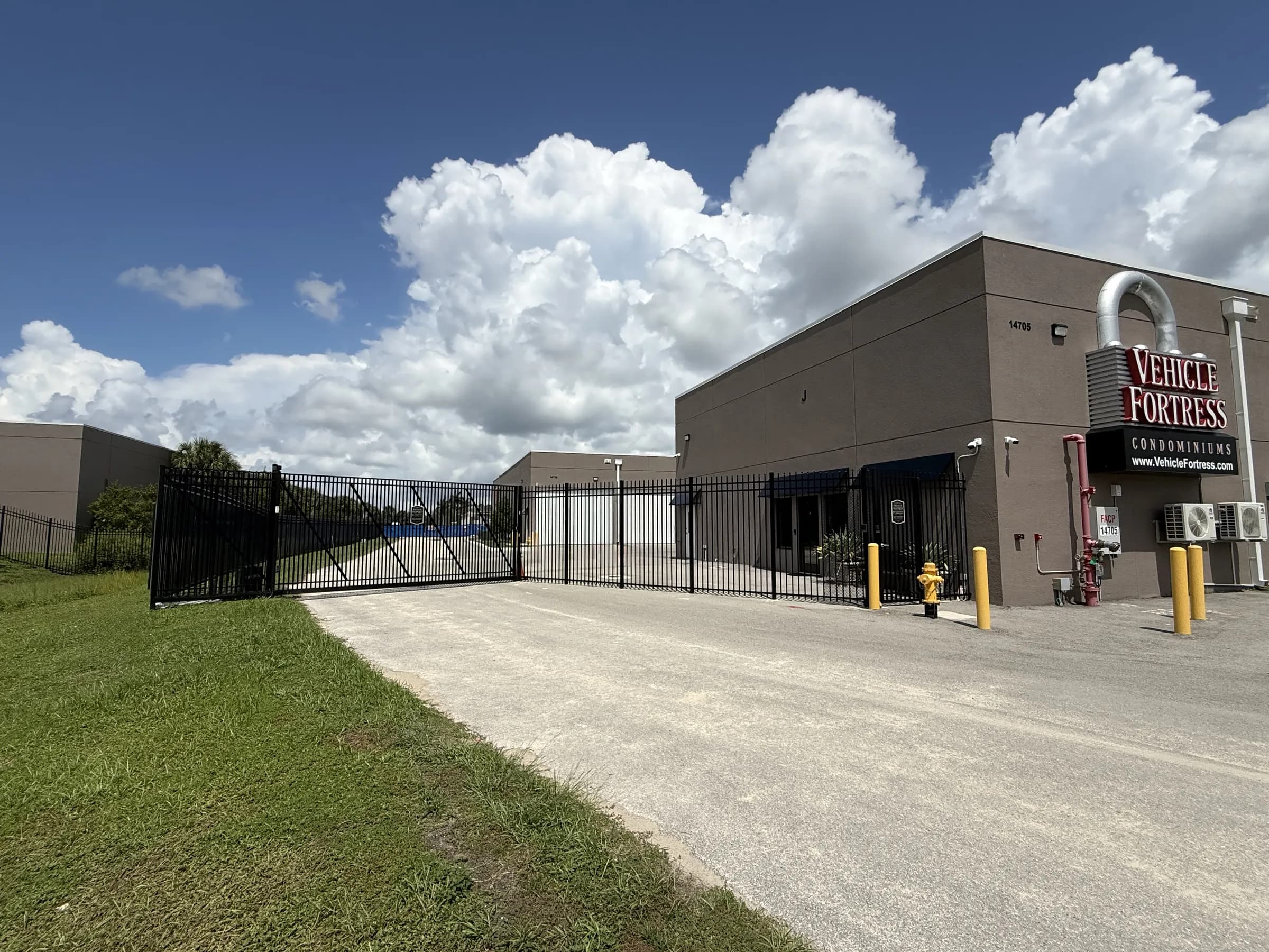 Angled view of the closed entry gate at left and the signed building corner at right, seen from the paved approach.