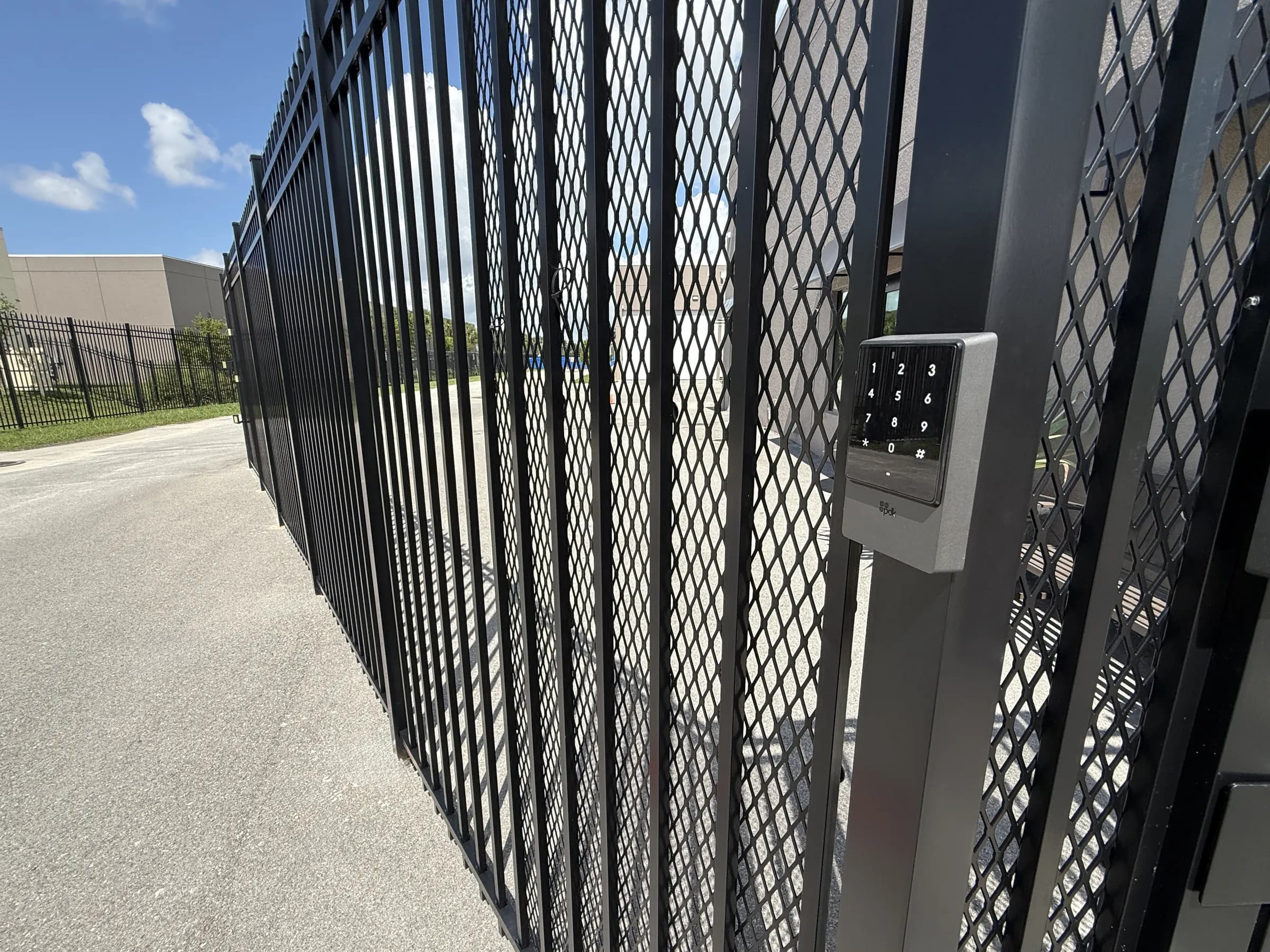Close-up of the illuminated numeric keypad on the black security gate post, fence line receding behind it.