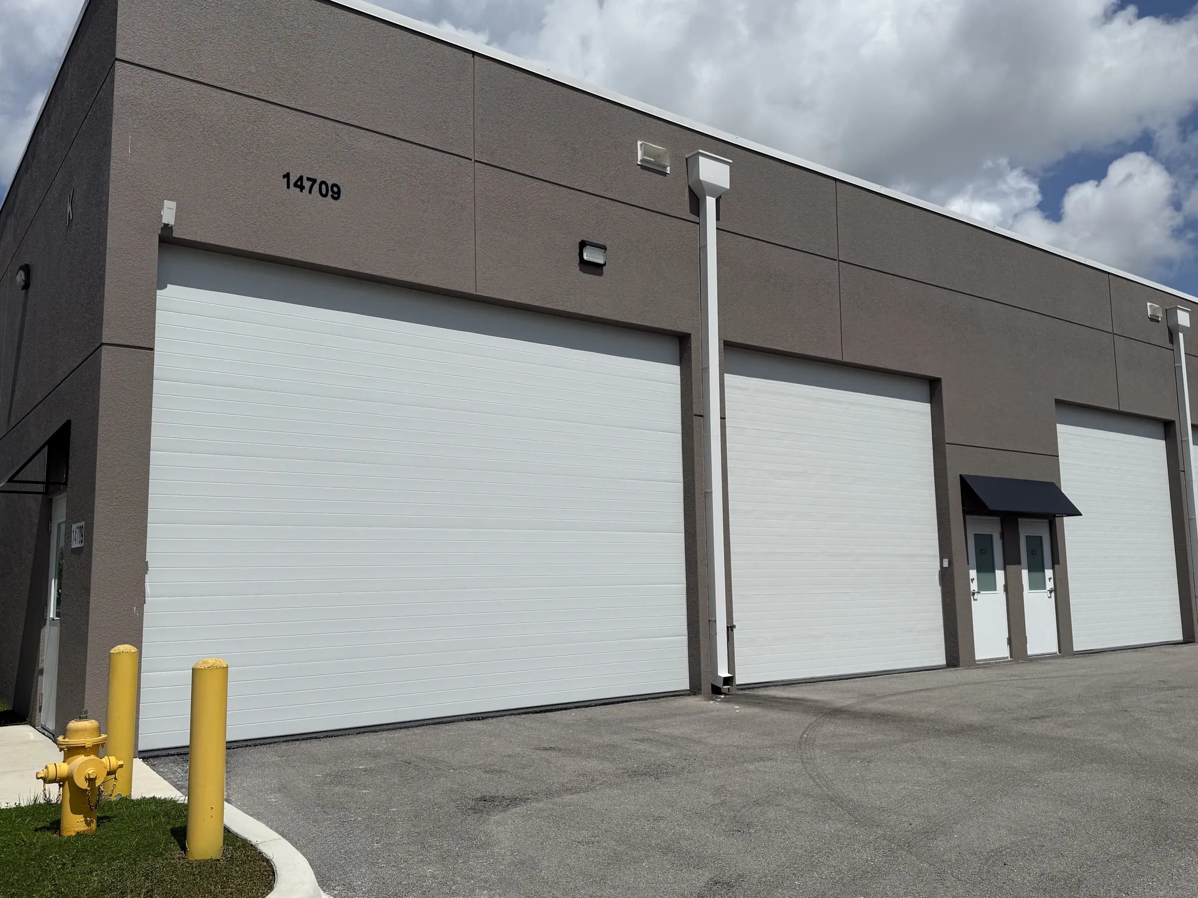 Three-quarter exterior of a taupe stucco building with three tall white sectional garage doors, entry doors under an awning, bollards and a fire hydrant.