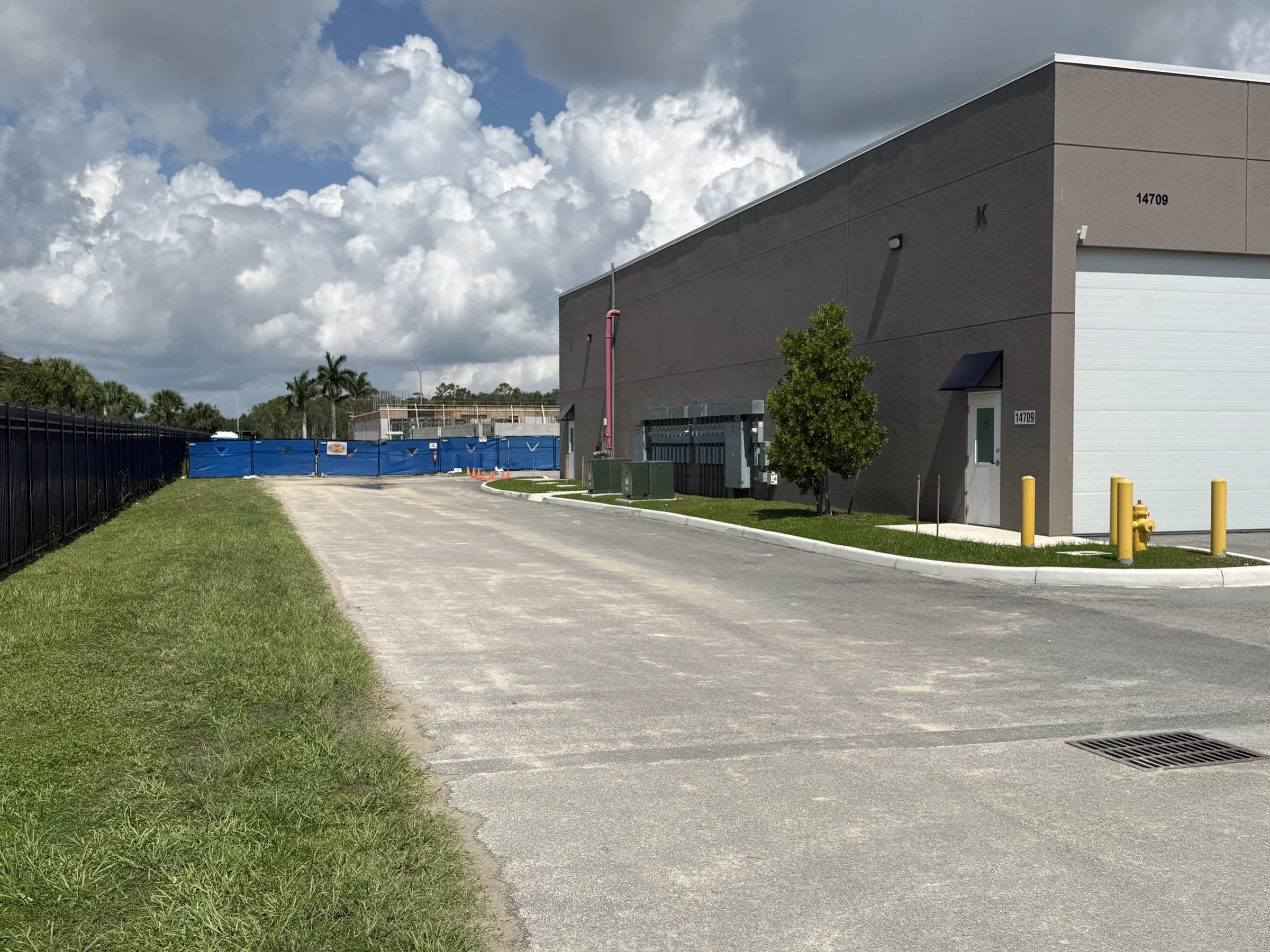 Ground-level view down a paved drive aisle past a building corner toward blue construction fencing and the active Phase B work area.