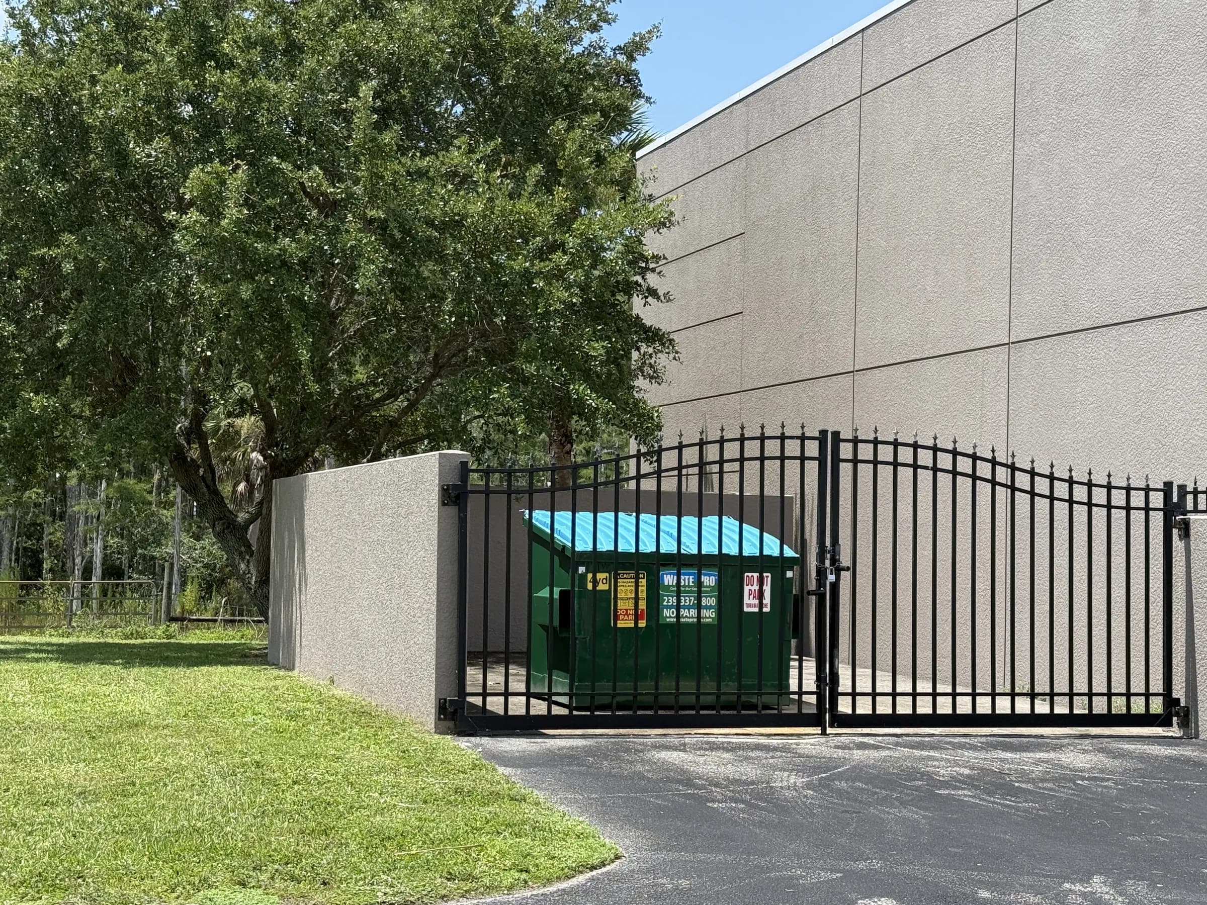 A black ornamental swing gate and stucco wall screening the dumpster beside a building wall, with a shade tree and lawn to the left.
