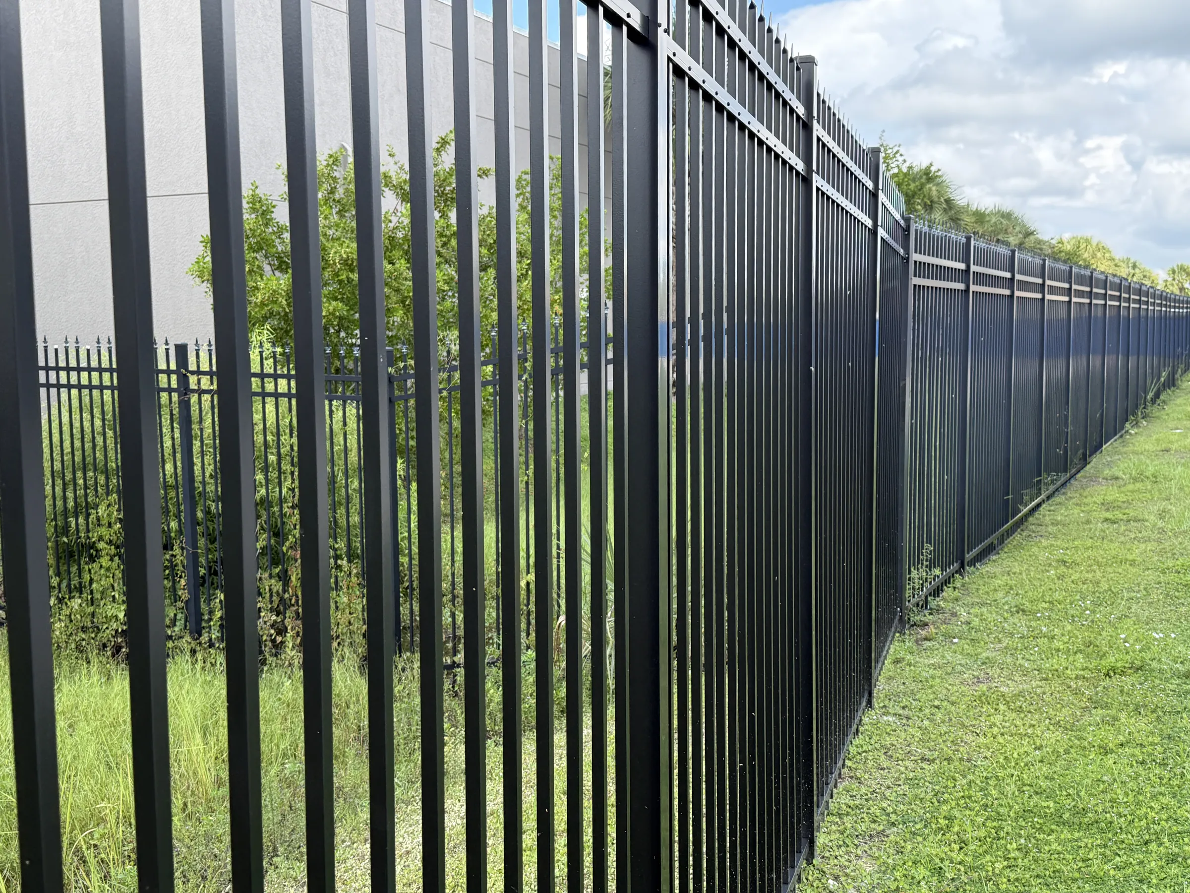 Low-angle view along the tall black aluminum picket perimeter fence receding over mowed grass, building wall and shrubs behind.