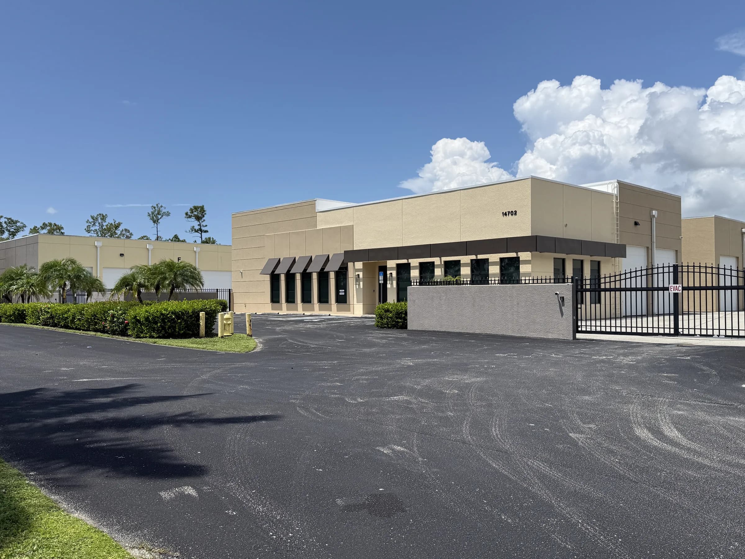 Wide view across the drive toward the office building's covered entry, with security gate, low hedges and palms under blue sky.