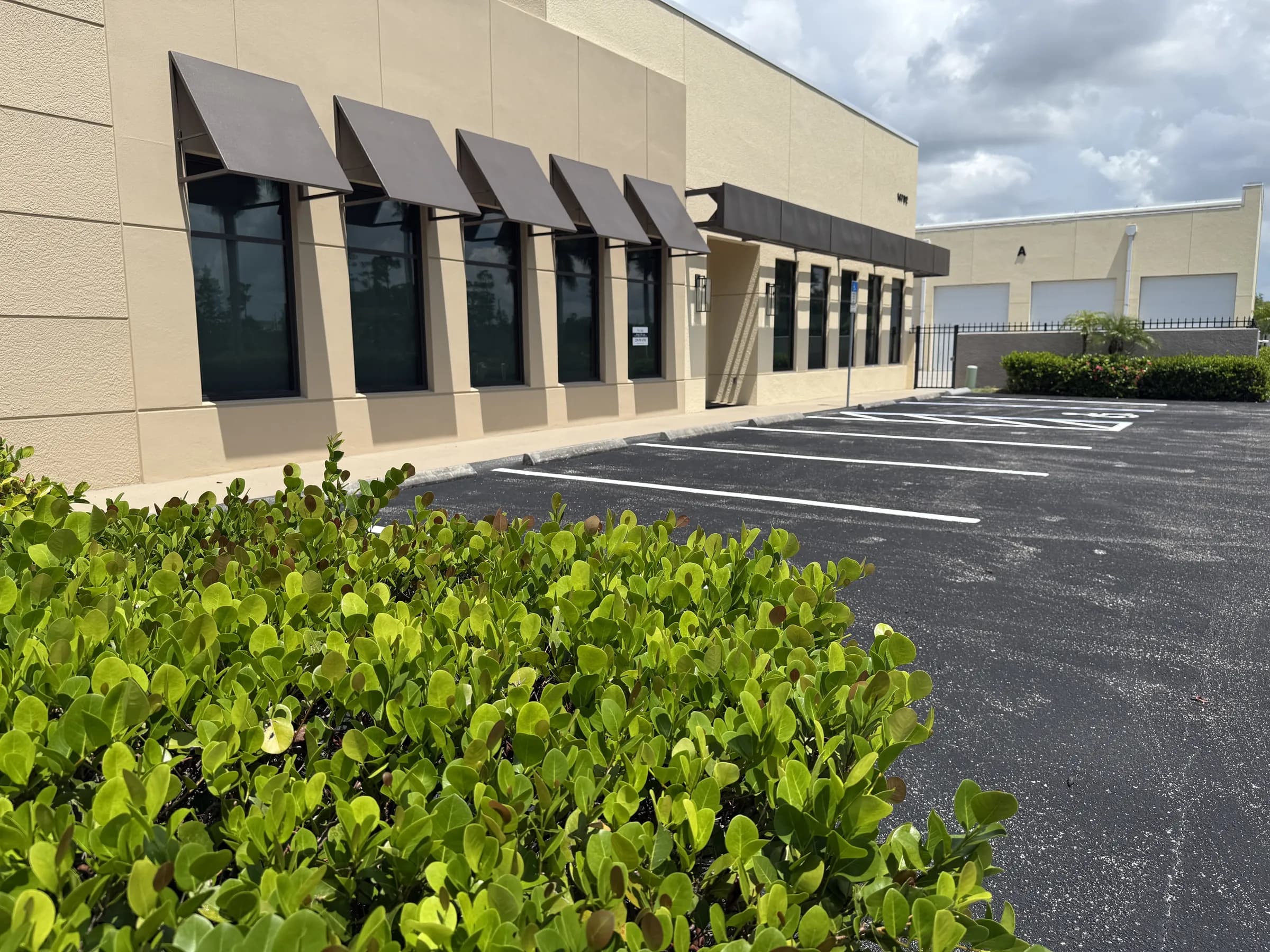 The office facade and striped lot seen past a green hedge, with the perimeter fence and storage bays behind.