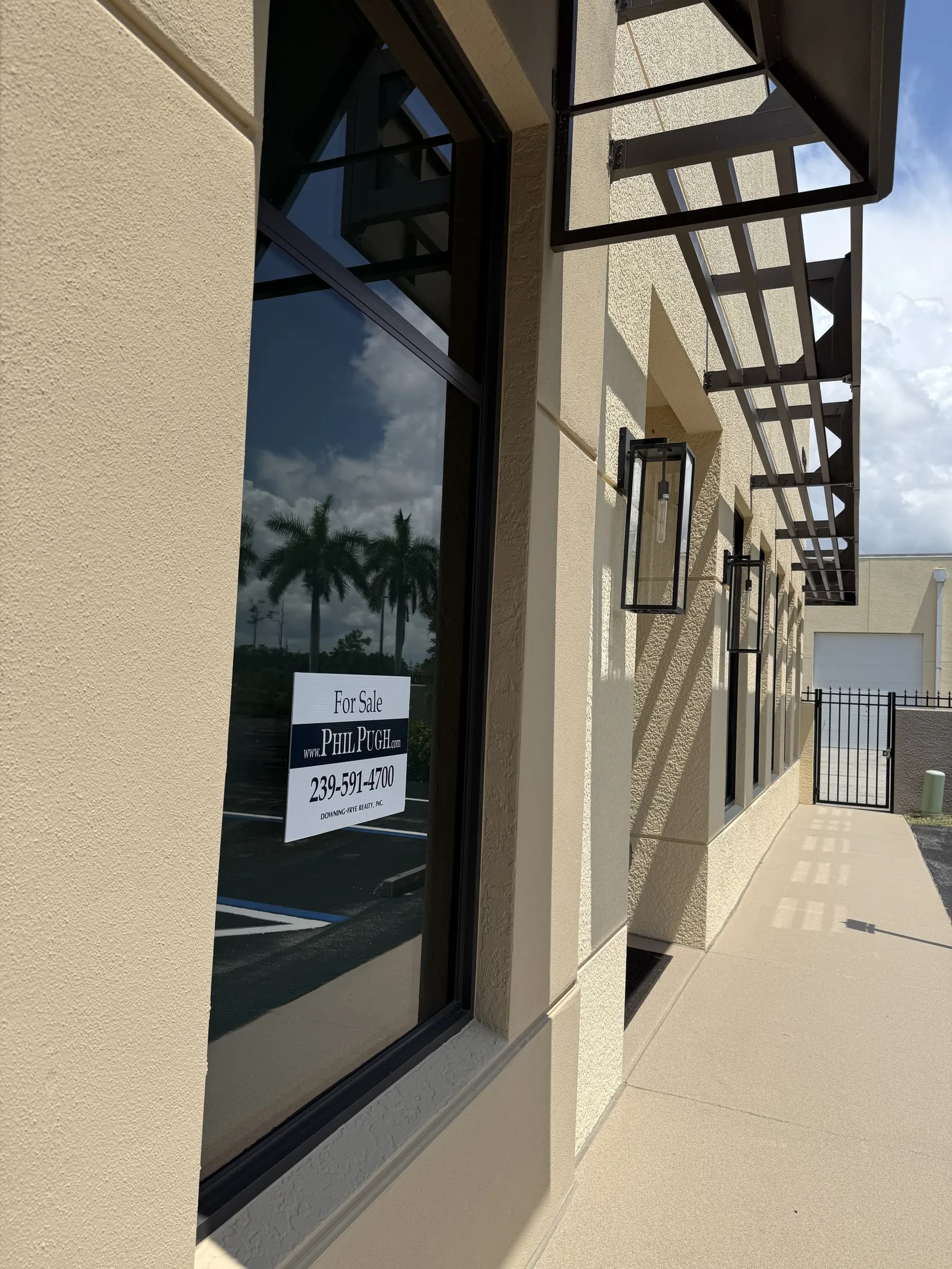 An office window reflecting palms and sky, with a Vehicle Fortress for-sale placard inside and awnings receding down the sunlit sidewalk.