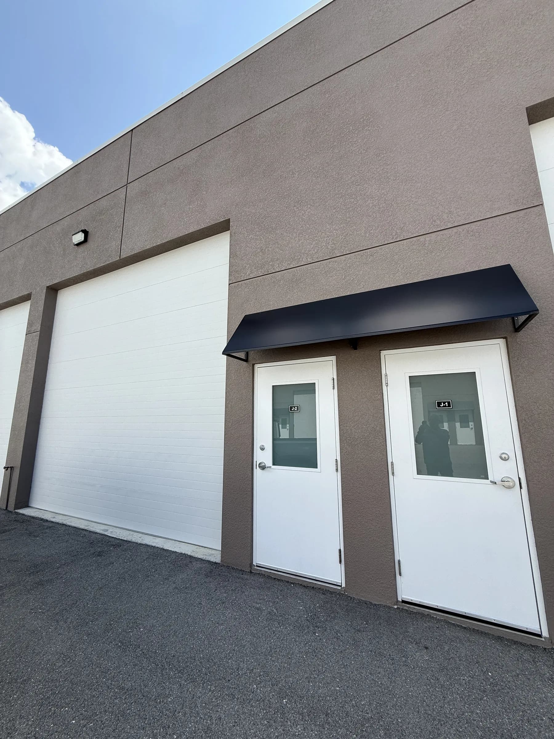 Low-angle exterior of two white side doors under a black awning beside a closed garage door on tan stucco, bright blue sky above.