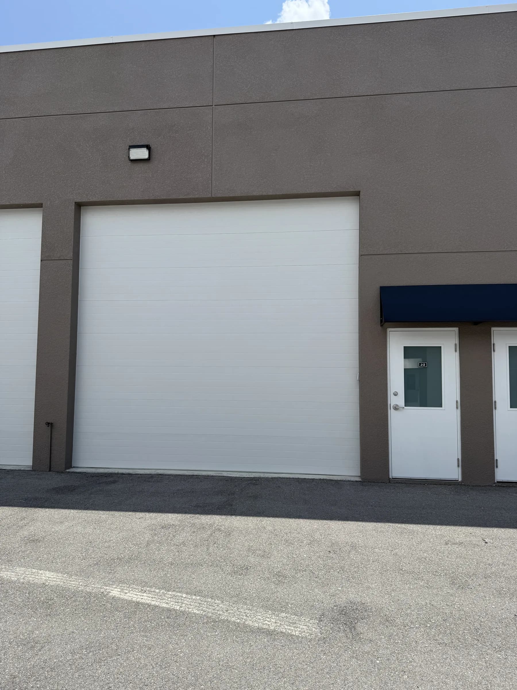 Exterior of a closed unit — tall white sectional garage door, wall-mounted light fixture and two white entry doors under a navy awning.