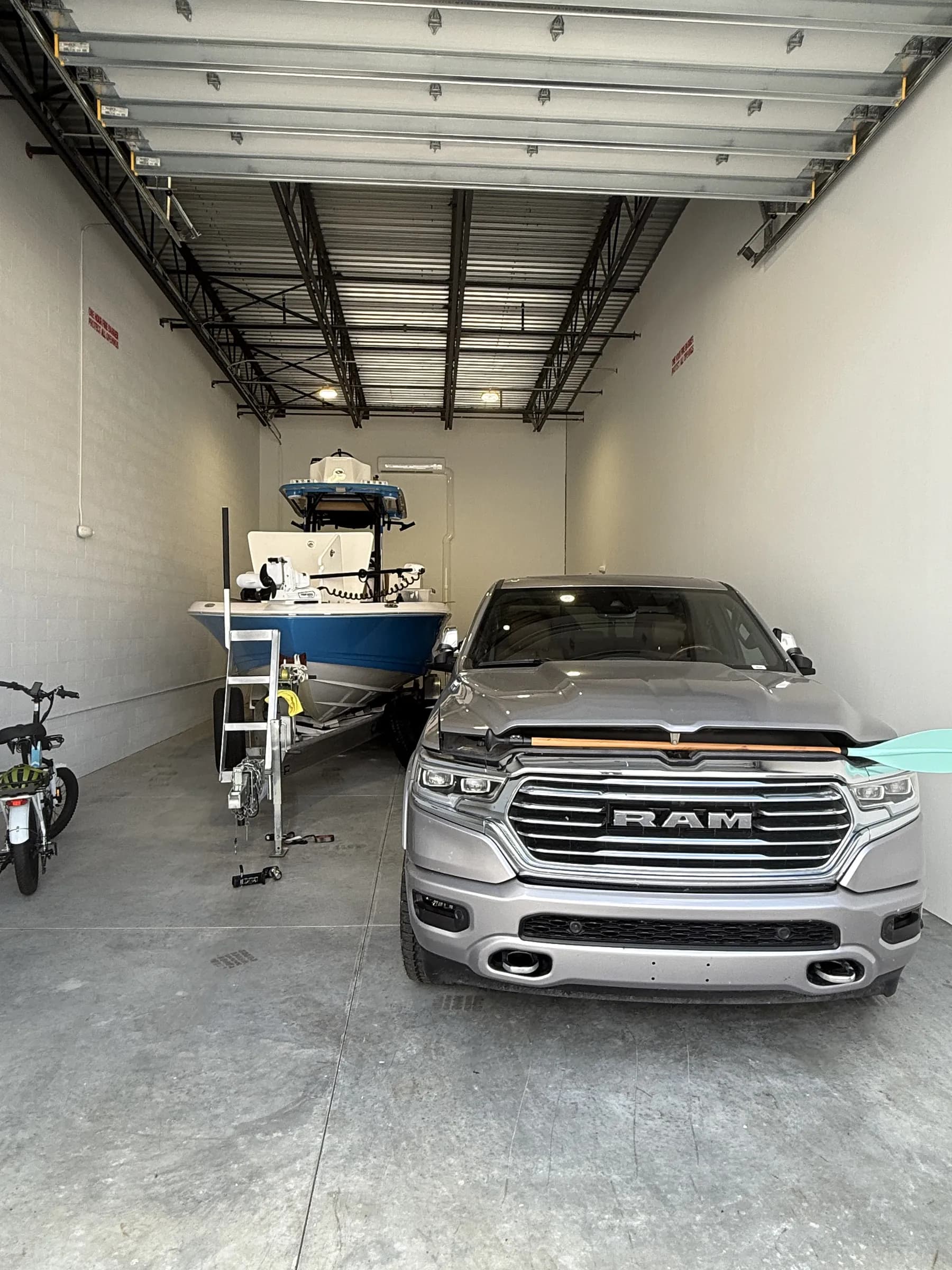 Interior from just inside the door — a pickup parked alongside a trailered blue-and-white center-console boat, bike at left and open trusses above.