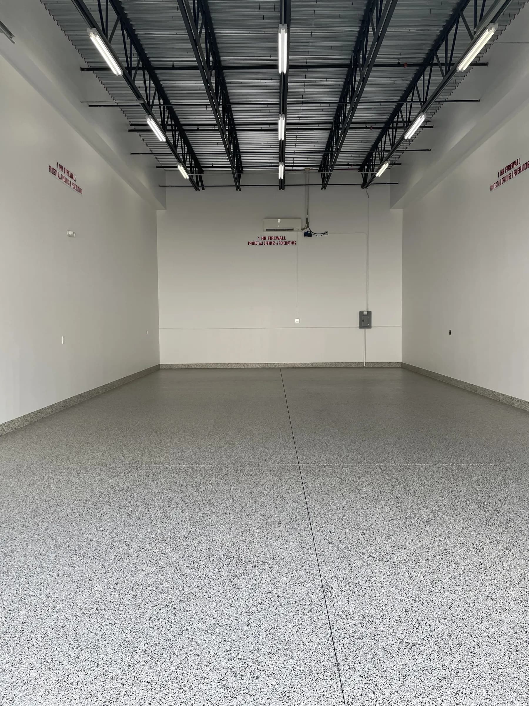 An empty finished unit seen from the door toward the rear wall — speckled epoxy floor, open steel truss ceiling, strip lighting and a wall-mounted mini-split.