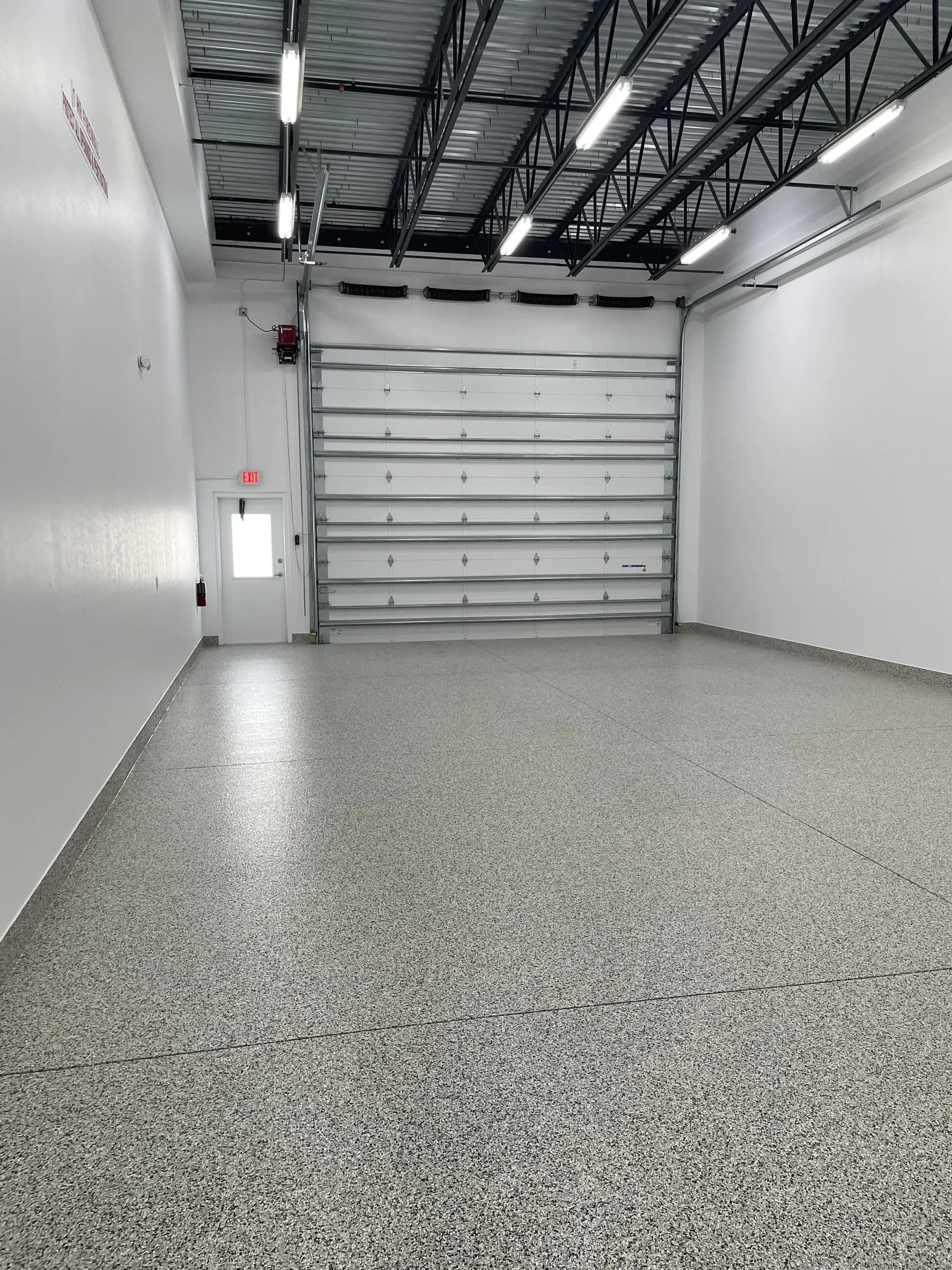 The same empty unit looking back toward the closed sectional garage door and side door, epoxy-flake floor and exposed trusses above.