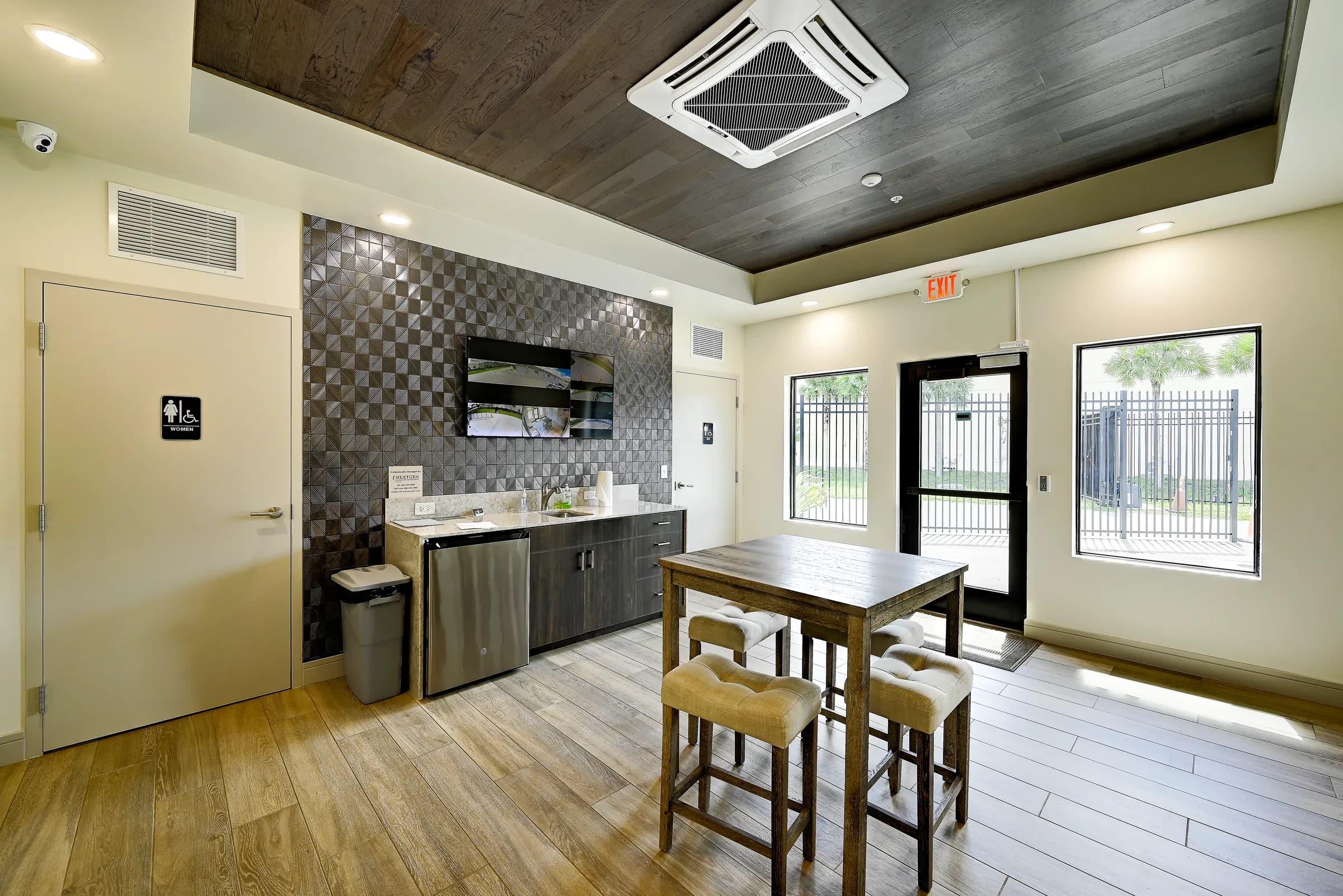 The common room lounge with kitchenette, geometric tile accent wall, TV, high-top table and the glass entry door.
