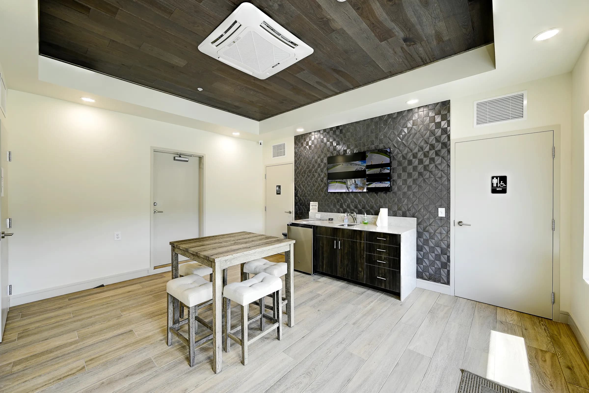 The common room kitchenette with dark cabinets, tile accent wall and TV, beside the restroom door.
