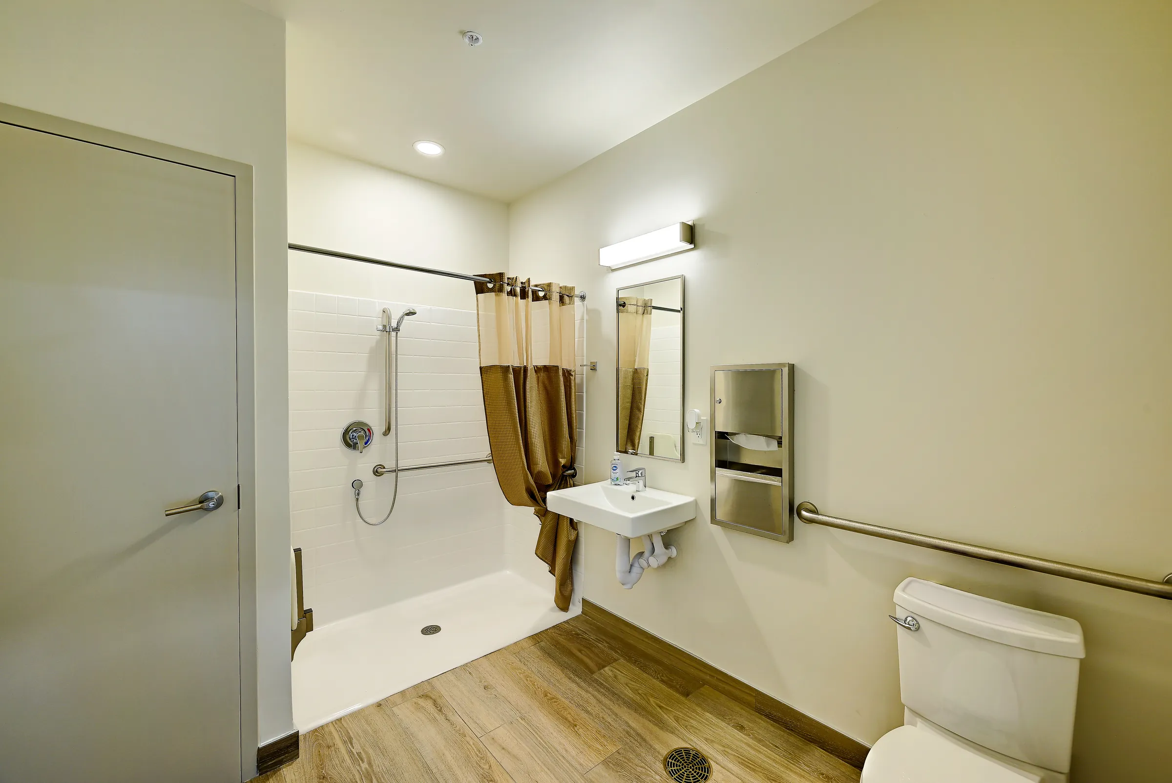 The common room's accessible restroom with a roll-in shower, grab bars, wall-hung sink and mirror.