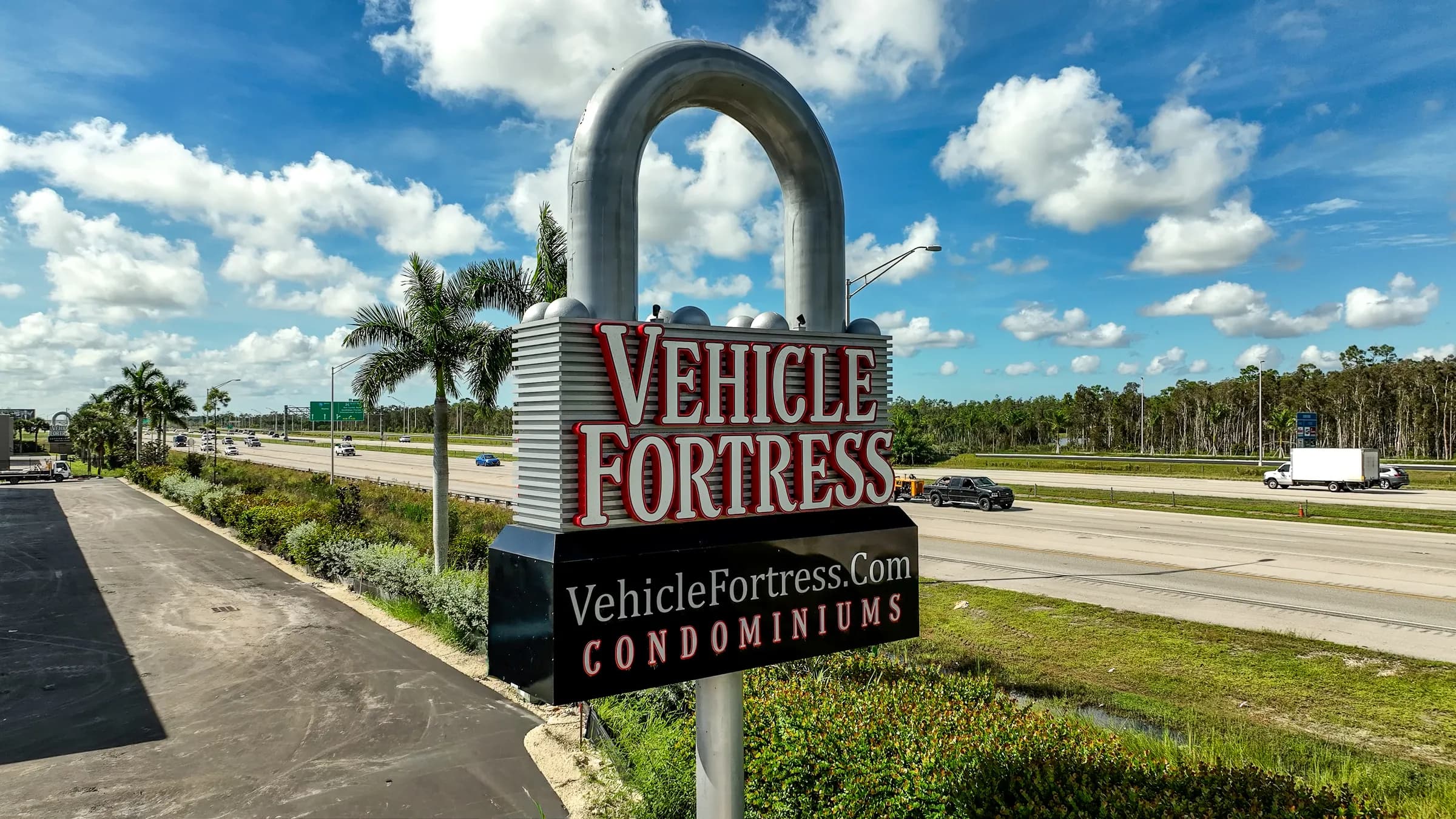 The padlock-shaped Vehicle Fortress Condominiums sign beside the highway, with palm trees and a bright blue sky.
