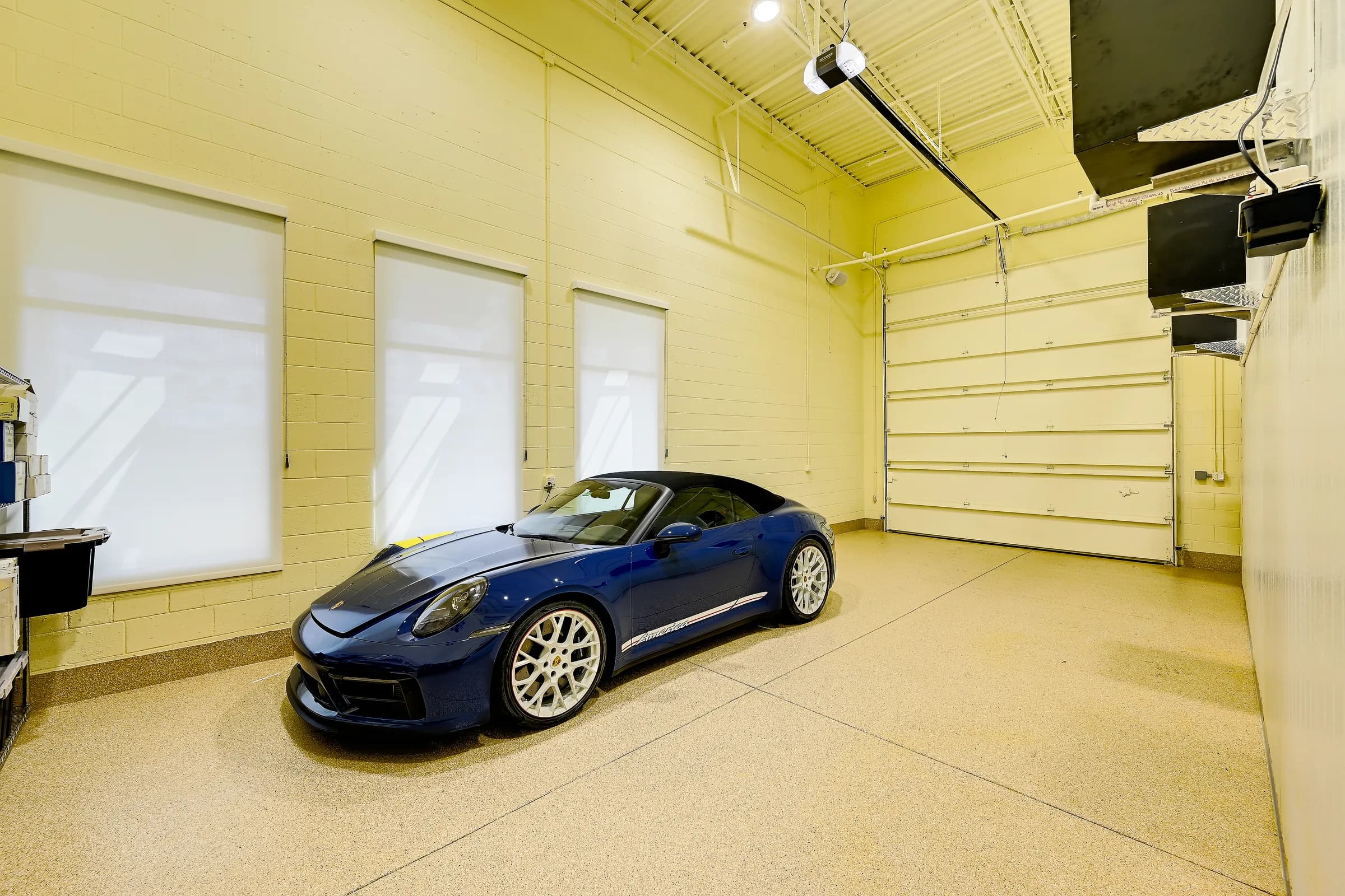 Front view of a blue convertible in the wine unit's garage, with window shades and the overhead door.
